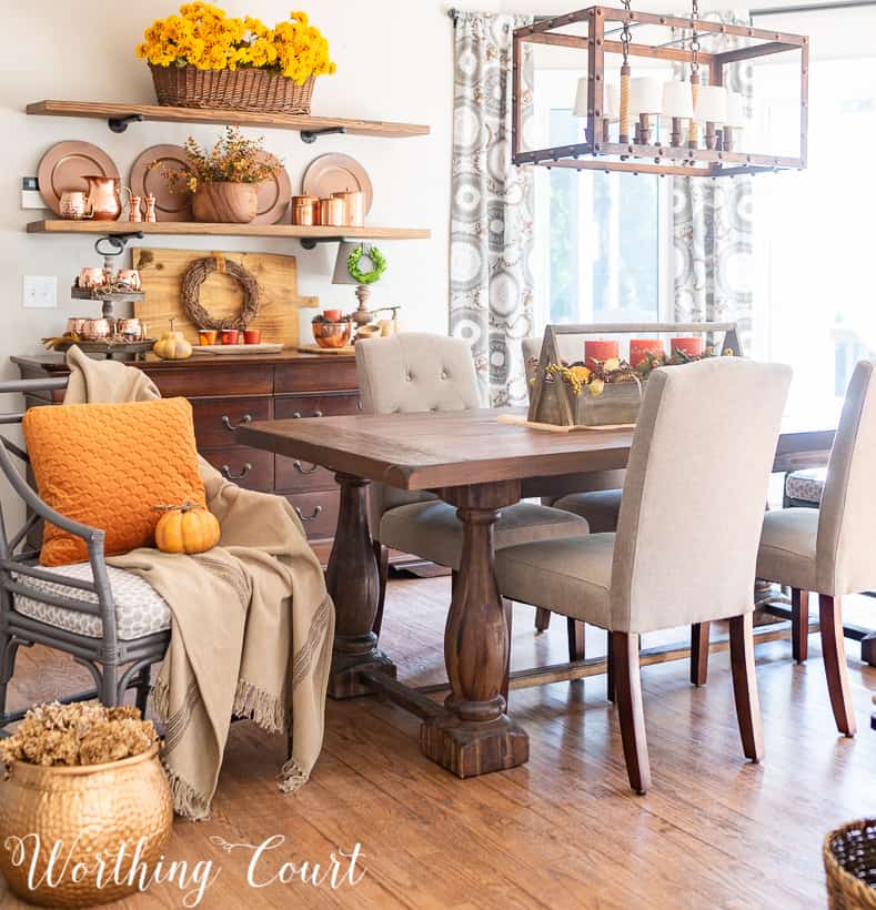 A large wooden table in the dining room with a chair and an orange pillow on the chair.   There is a rustic lighting fixture above the wooden table.