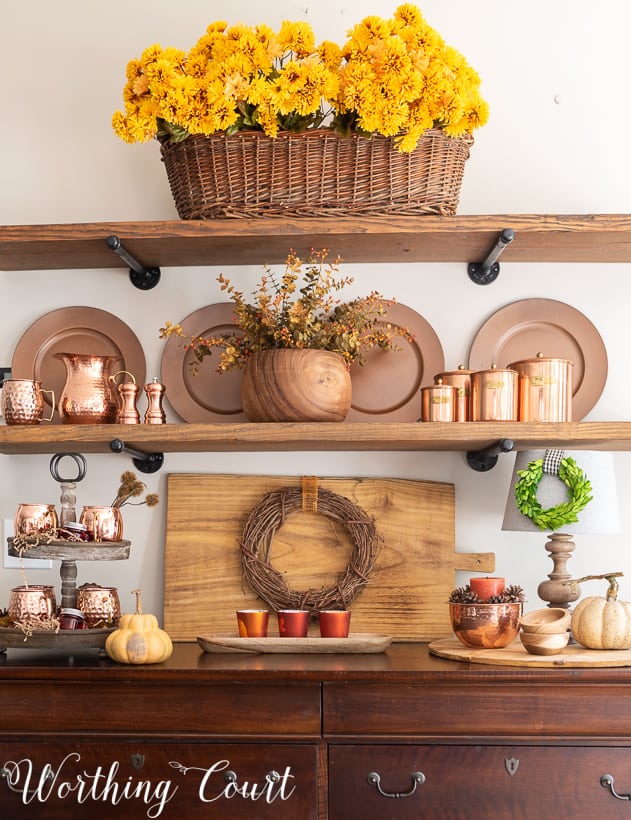 A large basket filled with Mum's, copper plates, copper cups and a wooden cutting board on the hutch.