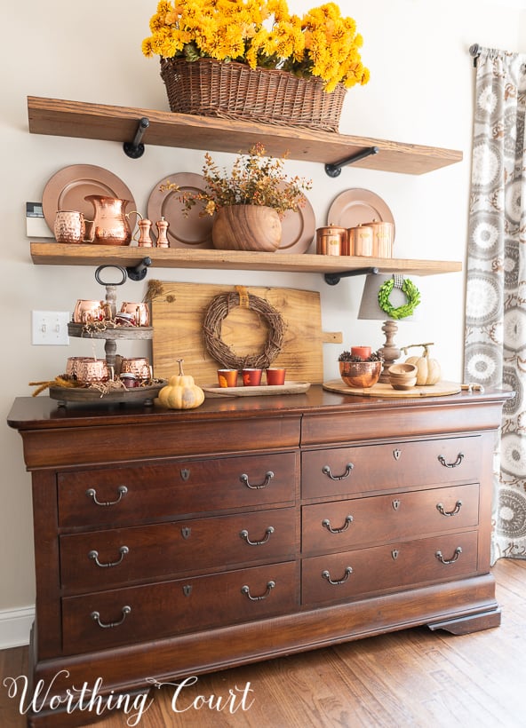 A wooden hutch with open shelves above it and copper accents on the shelves.   The top shelf has a a basket full of golden yellow flowers.