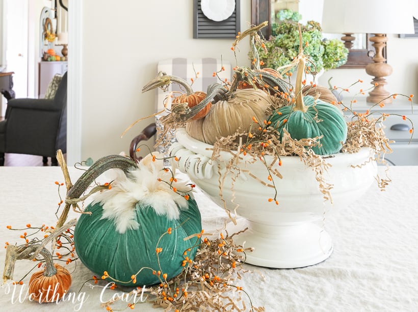 The velvet pumpkins are in a white ceramic bowl, with a large green and a small orange pumpkin beside it on the table.