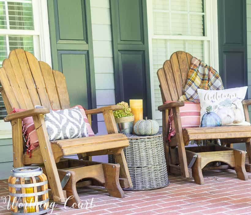 Two Adirondack chairs with fall pillows and a small pumpkin on a wicker table in between the chairs.