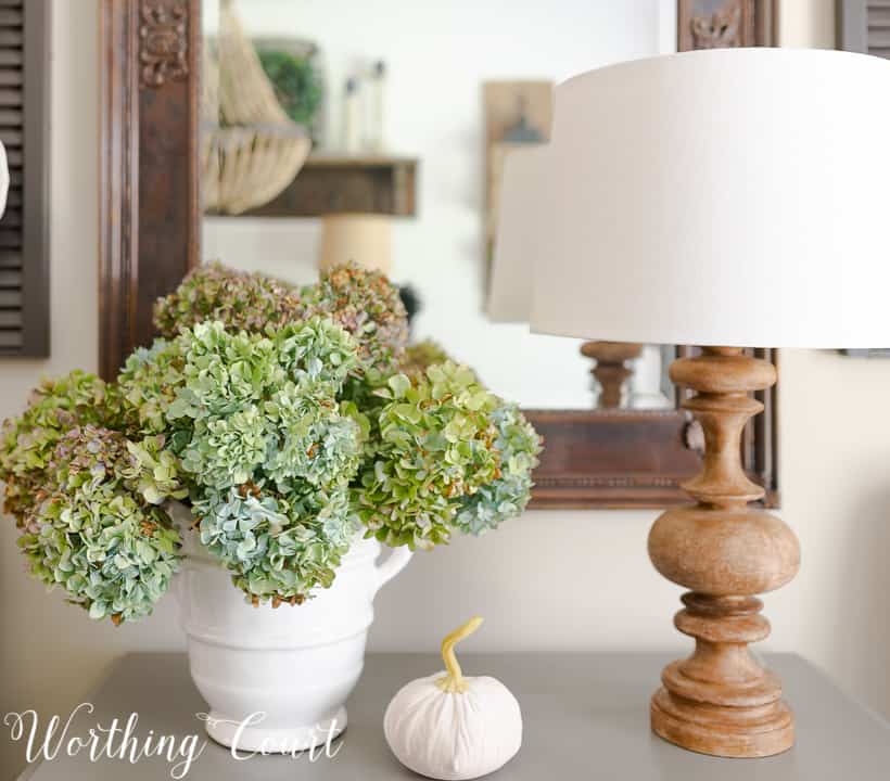 Blue hydrangeas in a white vase along with a small white pumpkin in between the flowers and a small lamp.