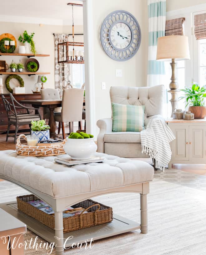 A tufted neutral ottoman in the living room with a large clock on the opposite wall.