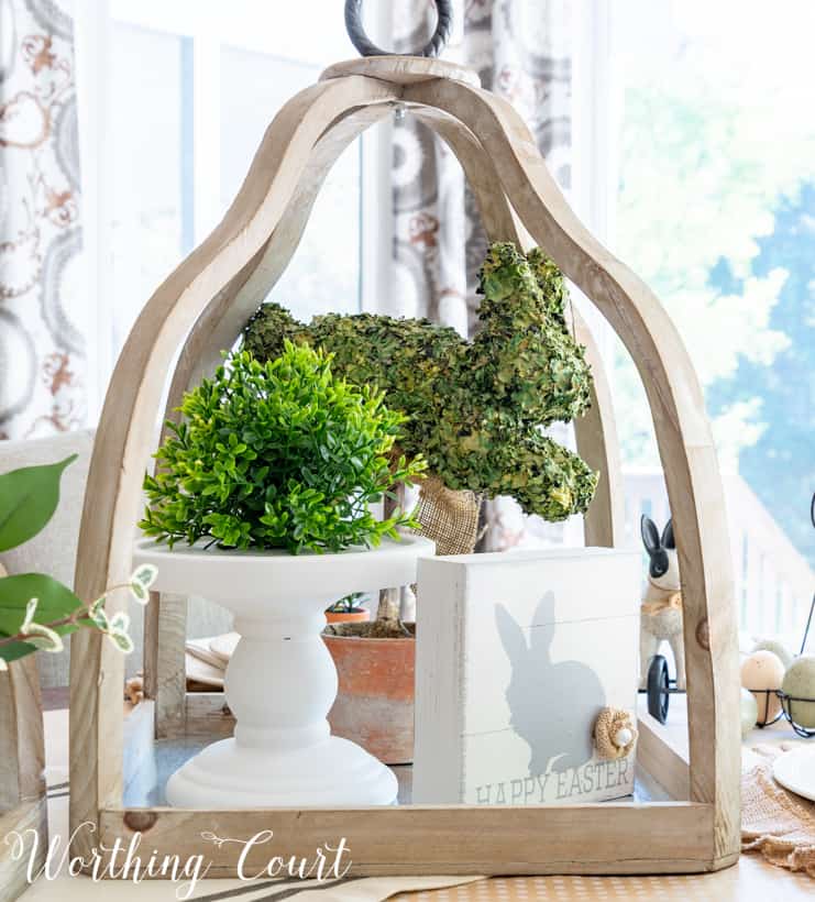 Large open lantern decorated for Easter with a white cake stand in it and a green potted plant on the stand.