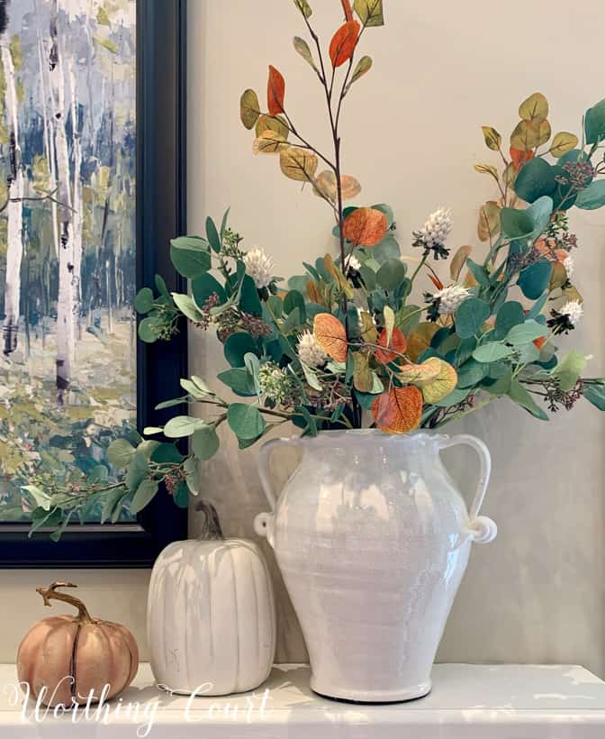 White ceramic urn filled with green and gold eucalyptus. There is a white pumpkin beside it and a smaller brushed copper one all on the mantel.