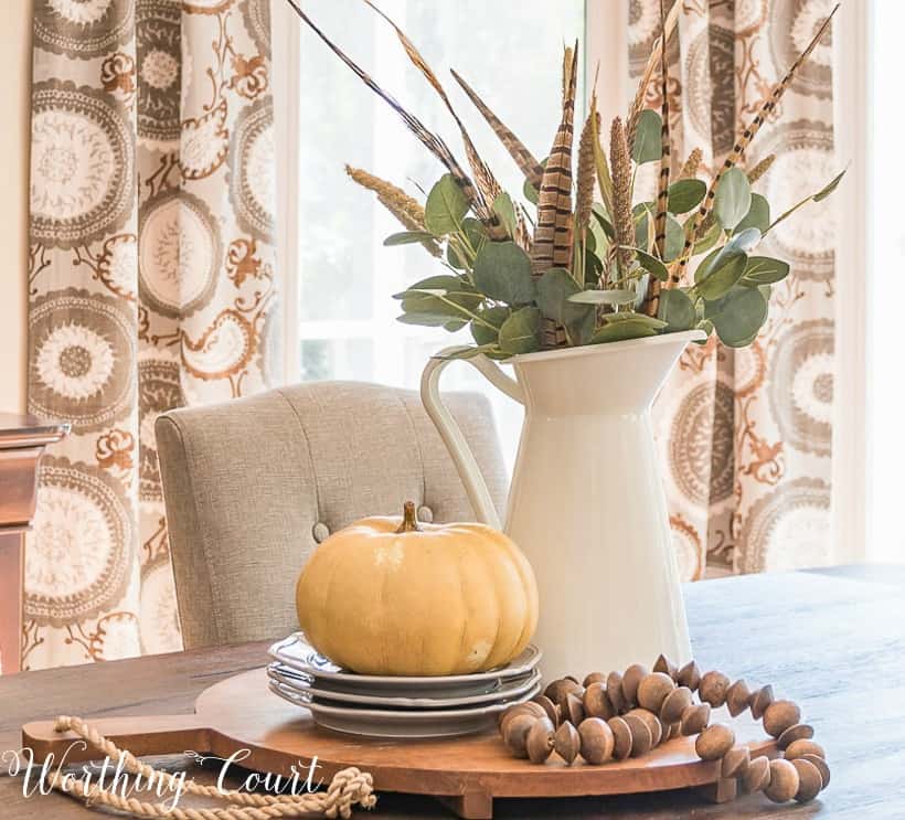 Fall centerpiece with wood beads and faux greenery in white pitcher. There is a small pumpkin placed on a stack of plates.