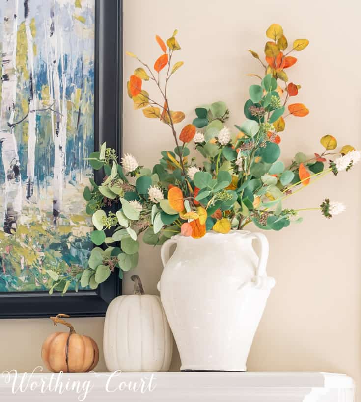 Faux fall eucalyptus stems in a white vase on a fireplace mantel.