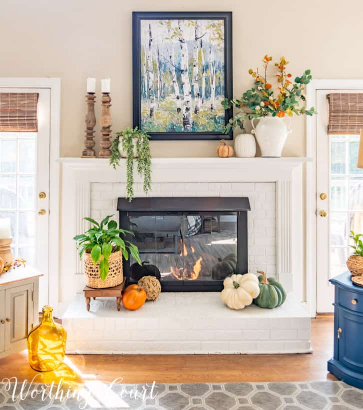 Brick fireplace painted white with fall decor such as small pumpkins in front of the lit fireplace.