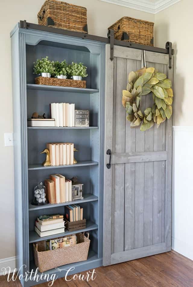 A wooden blue slate grey bookcase in the office.  It has a barn door style sliding door.
