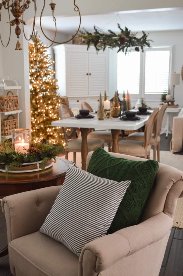 A neutral armchair with a green throw pillow on it. There is a lighted Christmas tree in the dining room.