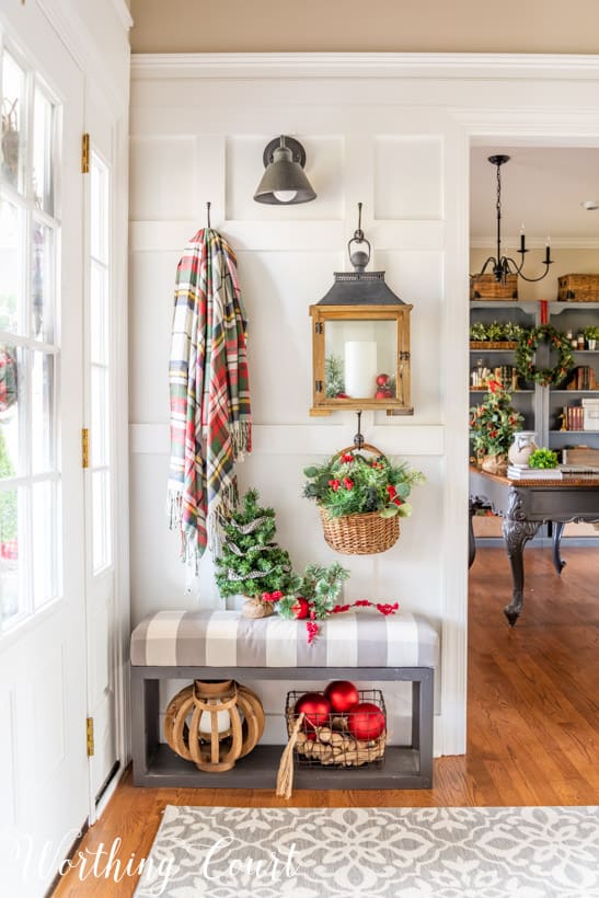 Board and batten wall with Christmas decorations hanging from hooks such as a plaid blanket and a small Christmas tree on the bench.