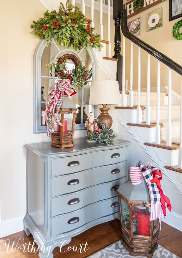 Small gray chest with an arched mirror above with a faux greenery swag and Christmas decorations.