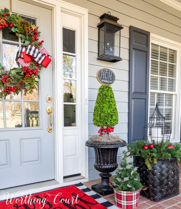 A mini Christmas tree is in a plaid container beside a black basket on the front porch.