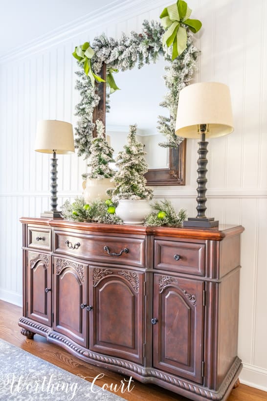 Sideboard with mirror above with flocked garland, flocked mini trees and green ribbon.