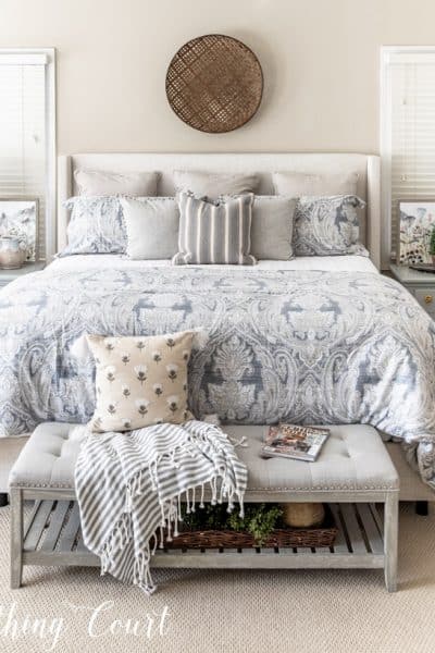 king size bed with upholstered headboard and blue, white and gray bedding with a bench at the foot of the bed.