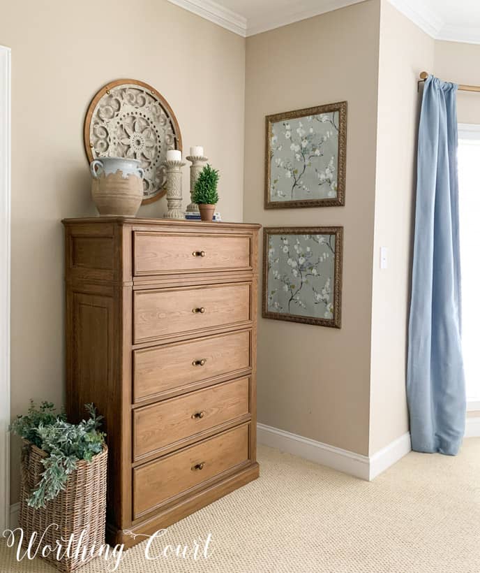 Primary bedroom chest of wooden drawers with blue draperies and framed wallpaper.