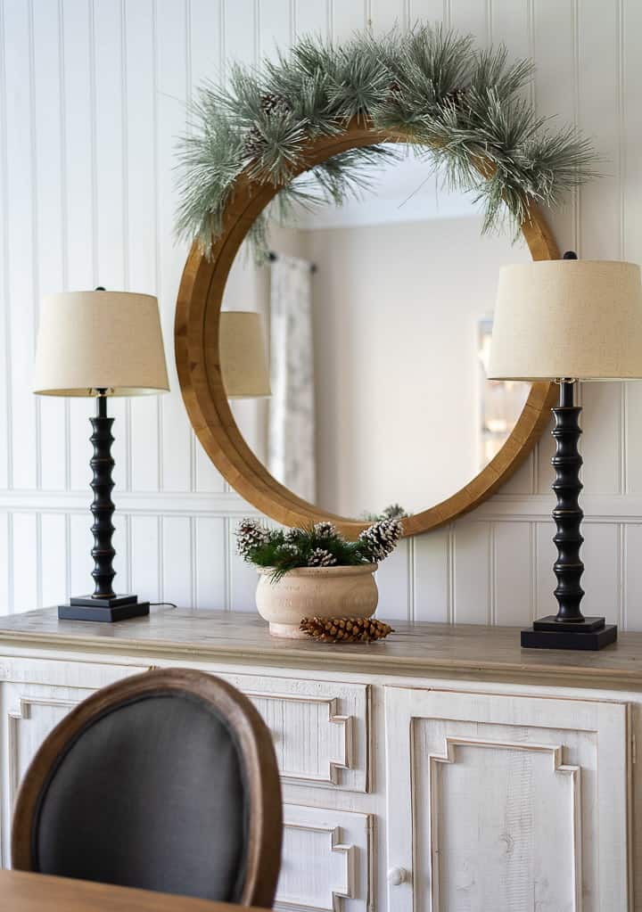 round mirror with greenery garland above over a white sideboard flanked by two lamps