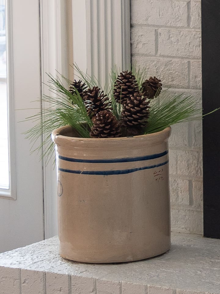 antique crock filled with faux pine greenery on a white fireplace hearth