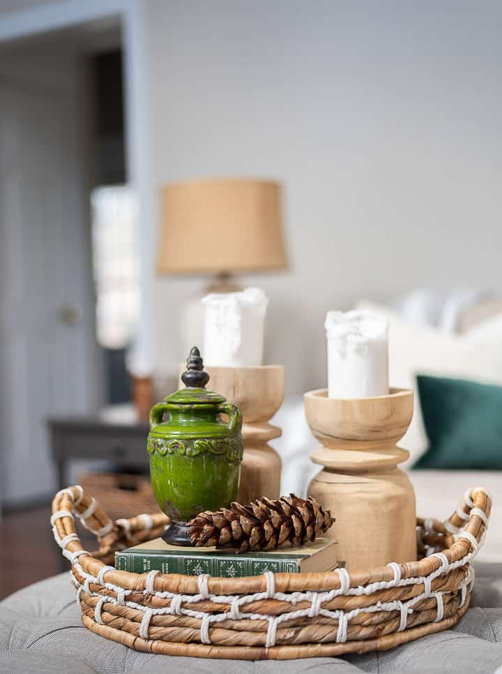 coffee table vignette in a round wicker tray basket with wood candlesticks, a green jar, a large pinecone and some stacked books