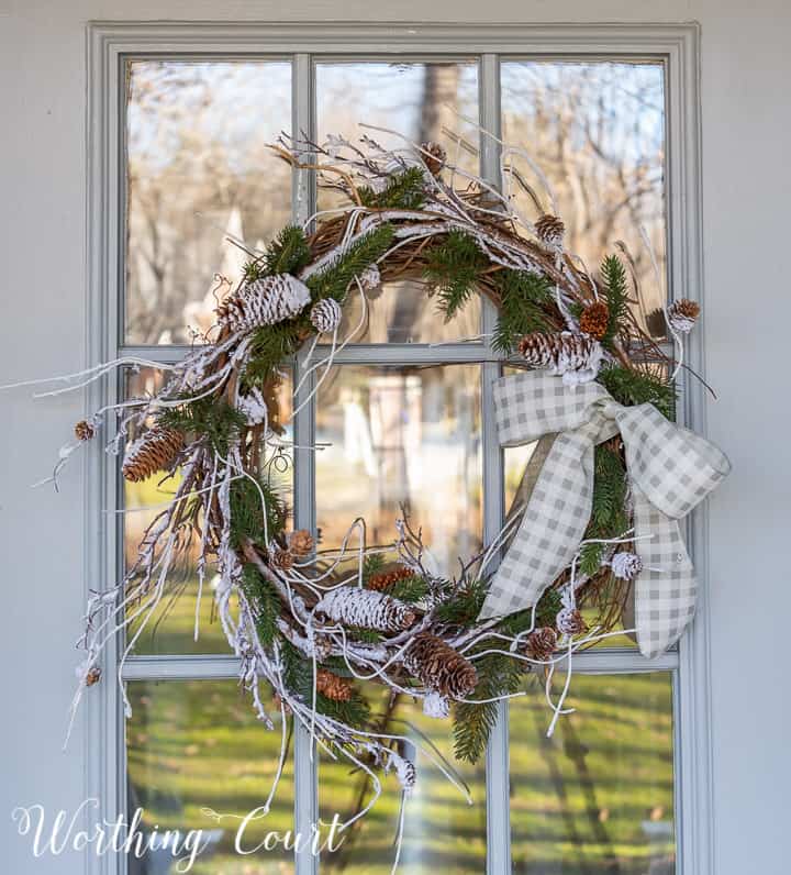 winter wreath with snow and pine branches and pinecones with a bow hanging on a glass door
