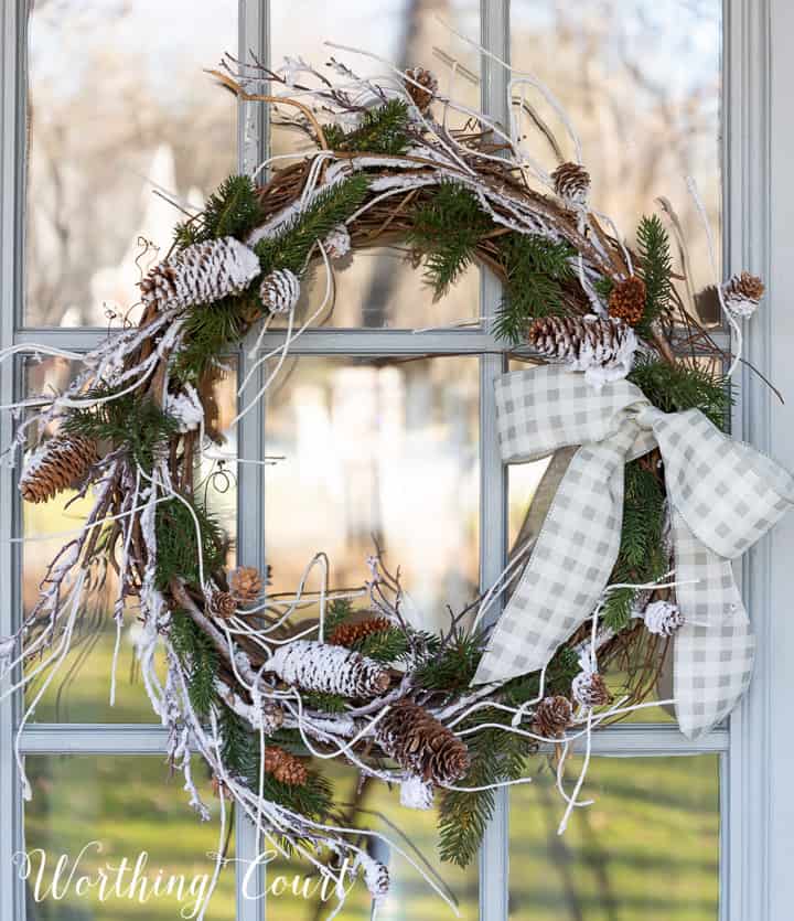 winter wreath with snow and pine branches and pinecones with a bow hanging on a glass door