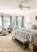 bay window with blue curtains, two side chairs and a king size bed with blue, white and gray bedding in a master bedroom