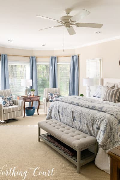 bay window with blue curtains, two side chairs and a king size bed with blue, white and gray bedding in a master bedroom