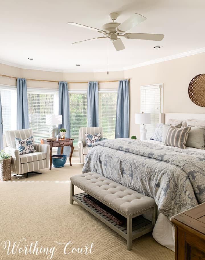 bay window with blue curtains, two side chairs and a king size bed with blue, white and gray bedding in a master bedroom
