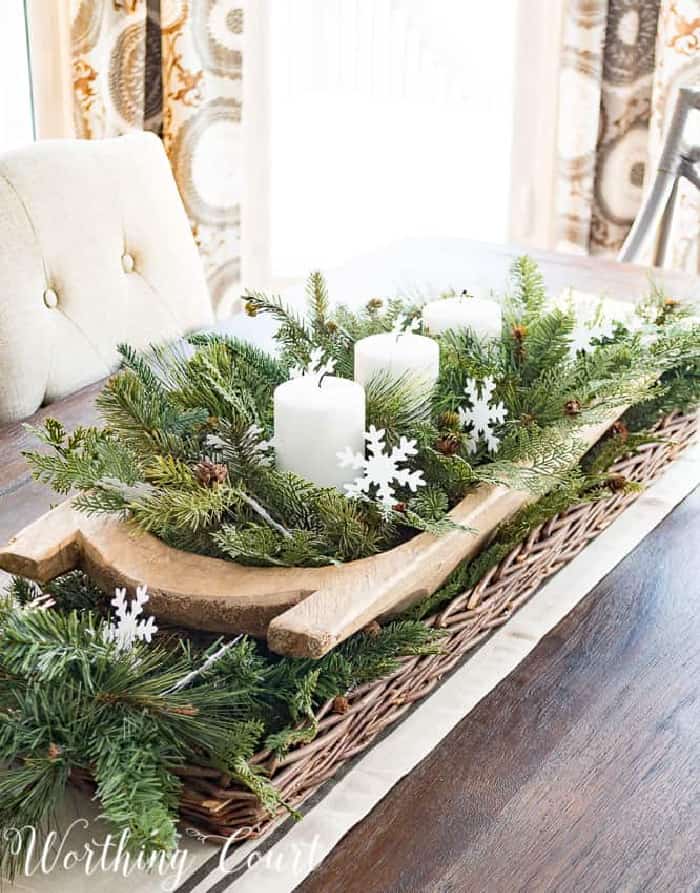 wood dough bowl nestled inside a long wicker basket filled with faux greenery, paper snowflakes and white candles