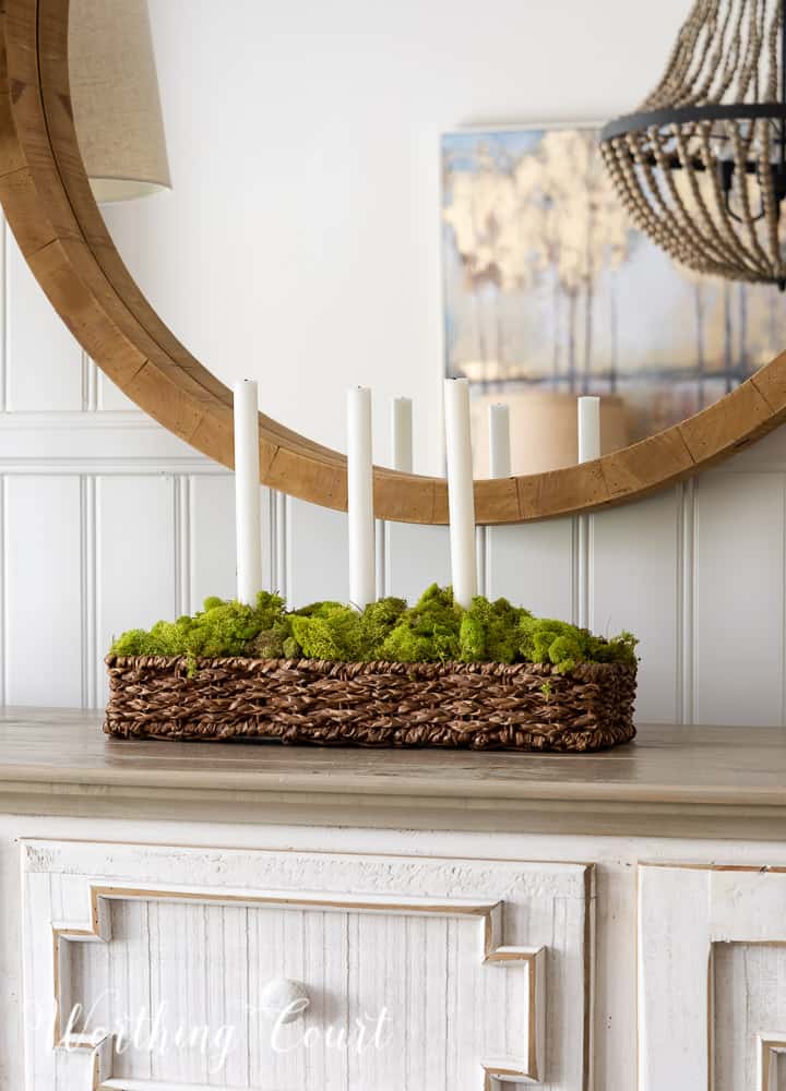 rectangular wicker basket with low sides filled with green moss and 3 tapered white candles on a sideboard