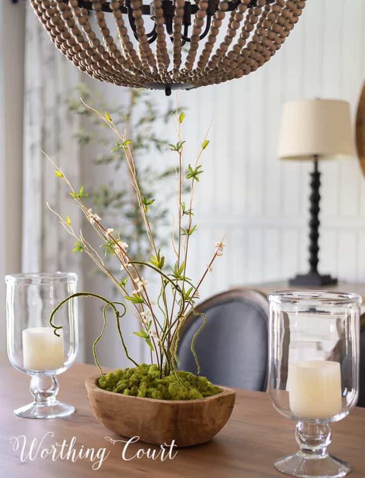 wood bowl filled with green moss and tall faux greenery flanked by a pair of glass hurricanes