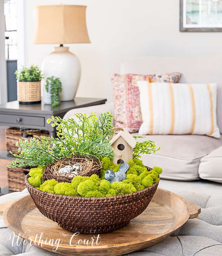 spring coffee table arrangement in a wicker bowl with moss, birdhouse, nest and greenery with couch in the background