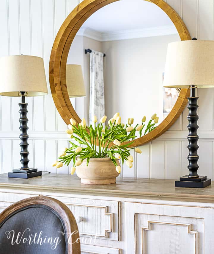 side board with round mirror above, two buffet lamps and white tulips in a wood bowl