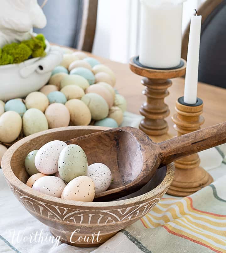 Partial view of Easter centerpiece with white ceramic bunny in the middle of an egg wreath and a wood bowl filled with faux Easter eggs