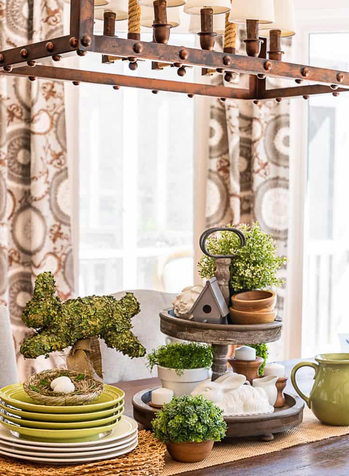 tiered tray decorated for spring with greenery, a birdhouse, candles and a bunny beside stacked plates and a bunny topiary