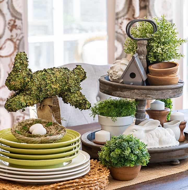 tiered tray decorated for spring with greenery, a birdhouse, candles and a bunny beside stacked plates and a bunny topiary