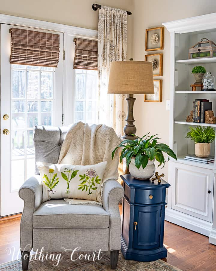 gray chair with tufted back with a floor lamp and blue side table with a spring pillow and live plant