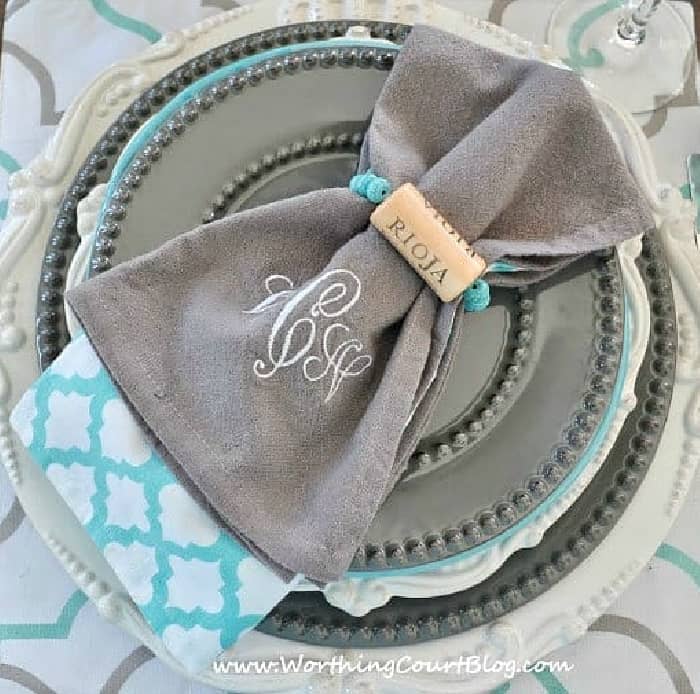 stack of gray and white plates topped with gray, white and blue napkins and a diy wine cork napkin ring