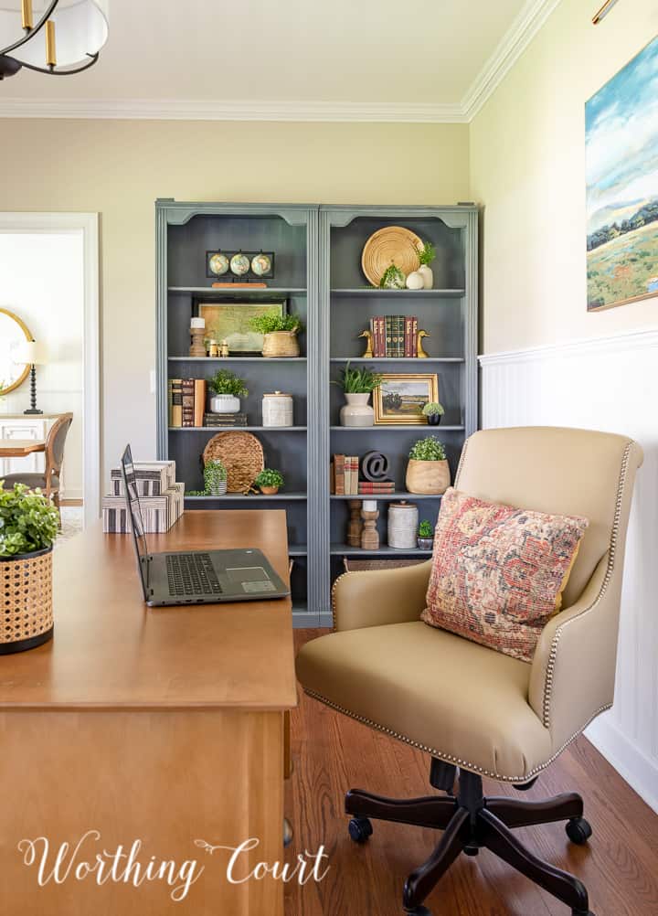 pair of gray bookcases filled with a variety of updated traditional accessories with a beige leather office chair in the foreground