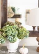 blue dried hydrangeas in a white urn on a gray chest in front of a mirror with a lamp and small white pumpkin