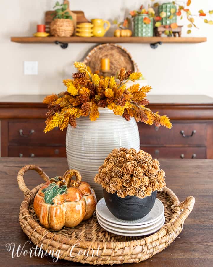 fall centerpiece in a woven tray with a containers filled with faux fall stems and a ceramic pumpkin box