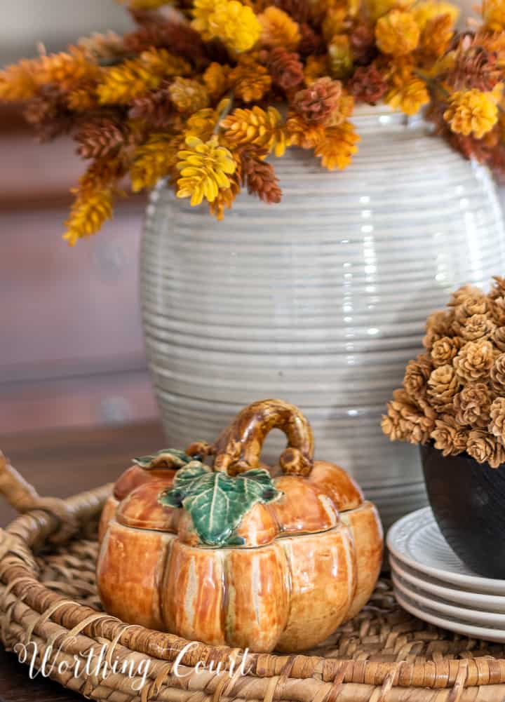 fall centerpiece in a woven tray with a containers filled with faux fall stems and a ceramic pumpkin box