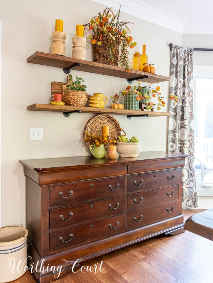 wooden open shelves above a sideboard decorated for fall with rich amber and green accessories