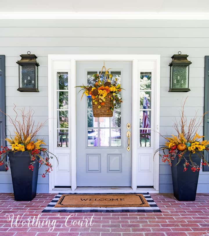 gray front door with a hanging basket and flanked with urns filled with faux fall stems and pumpkins in fall colors