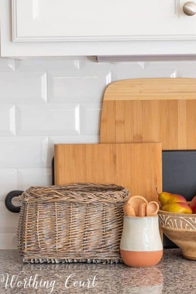 stacked wood cutting boards against a white subway tile backsplash with a basket, wooden bowl and small crock in front
