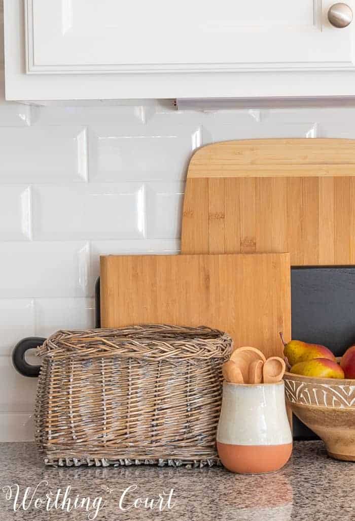 stacked wood cutting boards against a white subway tile backsplash with a basket, wooden bowl and small crock in front