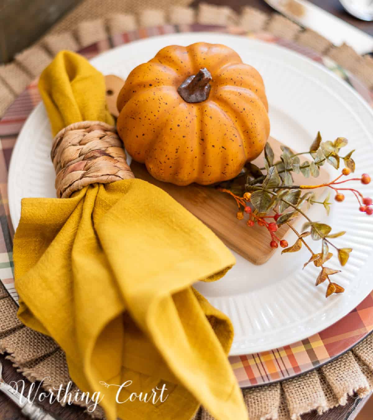 Thanksgiving place setting with a burlap placemat, plaid charger, white dinner plate and fall accessories.