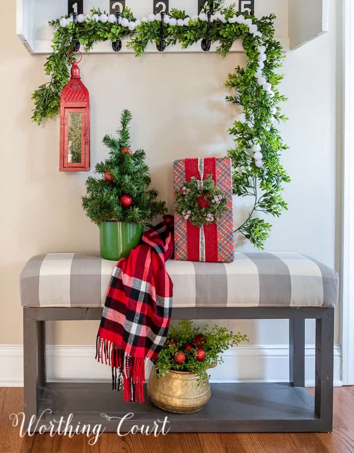 Gray bench with gray and white checked upholstery topped with Christmas decorations with a garland hanging on hooks above