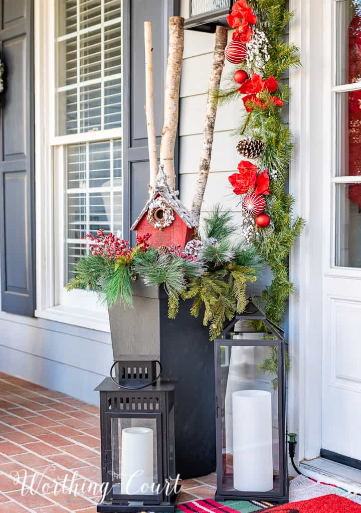front porch decorated for Christmas with red and white garland, planters, trees, lanterns, rugs and pillows
