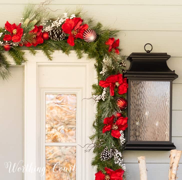front porch decorated for Christmas with red and white garland, planters, trees, lanterns, rugs and pillows